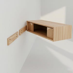 May include: A light-colored wooden floating shelf with a rectangular design. The shelf is mounted on a white wall, with two wooden mounting brackets visible. The shelf has an open front and sides.