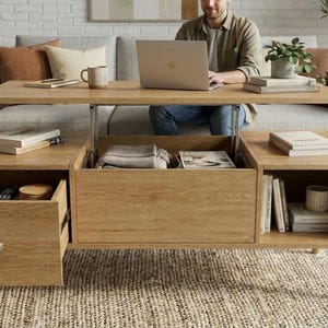 Puede incluir: Una mesa de centro de madera clara con tapa elevable y compartimentos de almacenamiento. Se encuentra frente a un sof&aacute; gris. La madera tiene un veteado natural. La mesa est&aacute; en un sal&oacute;n.