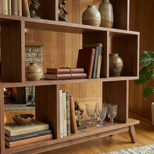 May include: A mid-century modern wooden bookshelf with multiple shelves. The shelves display books, decorative vases, and small sculptures. A fireplace with a burning fire is visible in the background, along with a potted plant.