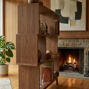 May include: A mid-century modern style wooden bookshelf with multiple open shelves. The bookshelf is filled with books, vases, and decorative objects. The wood has a rich, warm tone. The bookshelf is in a living room setting with a fireplace and artwork.
