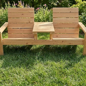 May include: A light brown wooden bench featuring two separate seating areas with horizontal slat backrests and a central square table surface connecting the two seats, supported by four legs and two armrests on the outer sides of the structure.