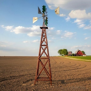 May include: A rustic wooden windmill stands tall in a cultivated field under a blue sky with fluffy clouds. The windmill has a brown frame and silver and green blades. In the distance, a red barn and house are visible.