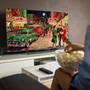 May include: A flat-screen TV displays a vintage Christmas scene with classic cars and people. The image on the TV shows a street with holiday decorations, including "Merry Christmas" and "Happy Christmas" signs. A person holds a bowl of popcorn.