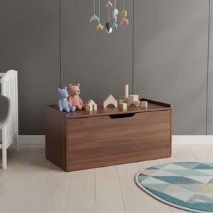May include: A dark brown wooden toy chest with a recessed handle. The chest is rectangular. Two teddy bears and wooden blocks are on top. A colourful mobile hangs above. A round rug with geometric patterns is on the floor.