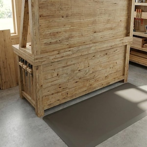 May include: A large, wooden workbench with a natural finish, featuring a built-in tool rack and a gray anti-fatigue mat on the floor. The workbench has a sturdy, rustic design, ideal for woodworking projects.