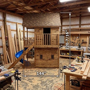 May include: A wooden playhouse with a brown shingled roof, a blue slide, and a balcony. The playhouse is 144 inches wide and 220-7/16 inches tall. The playhouse has a rustic, natural wood finish and is set in a workshop.