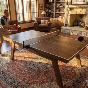 May include: A wooden ping pong table with a net, set up in a living room. Two people are playing table tennis. The table is brown with a white border. The room has a fireplace, a piano, and leather chairs.