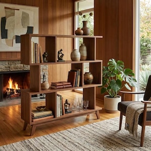 May include: A mid-century modern wooden bookshelf with multiple cubby shelves. The shelves are filled with books, vases, and decorative objects. A fireplace and a large houseplant are visible in the background.