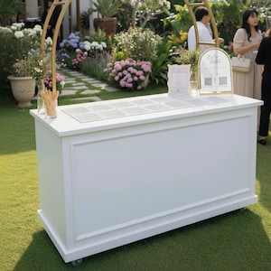 Puede incluir: Una barra rectangular blanca con un panel decorativo y ruedas, colocada sobre un césped verde. La barra tiene un arco dorado encima, con arreglos florales y señalización. El fondo presenta un jardín con flores y personas.