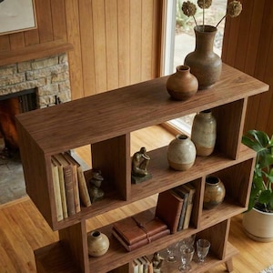 May include: A dark wood bookshelf with multiple shelves, displaying books, decorative vases, and small sculptures. The bookshelf is in a room with a fireplace and a large window, creating a warm and inviting atmosphere.