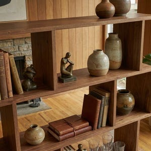 May include: A wooden bookshelf with a unique, geometric design. The shelves display various items, including books, ceramic vases in shades of brown and beige, and small sculptures. The warm wood tones create a cozy, inviting atmosphere.