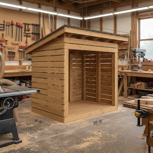 May include: A wooden shed with a sloped roof and open front. The shed is constructed from horizontal wooden planks and has dimensions of 2.44 metres by 3.05 metres. The roof is made of corrugated metal. The shed is in a workshop setting.