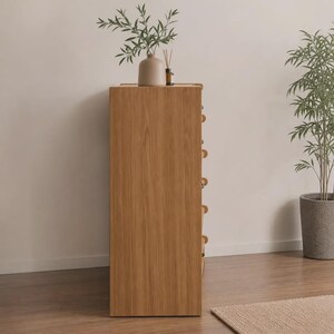 May include: A tall, rectangular wooden chest of drawers with a light oak finish, featuring a smooth side panel with visible wood grain. The unit has multiple drawers stacked vertically, each equipped with small, round, light-colored wooden knobs.