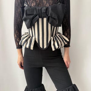 Peut inclure: Un corset à rayures noires et blanches avec un grand nœud noir. Le corset a un basque à volants et est associé à un pantalon noir à ourlets à volants. Le haut est en dentelle noire.