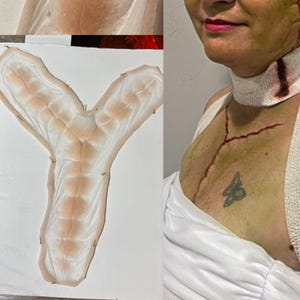 May include: A prosthetic Y-shaped wound appliance with a flesh-toned base and detailed vein-like patterns. The image also shows a person with makeup simulating a neck wound and chest lacerations, with a small tattoo.