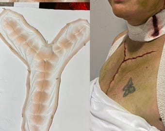 May include: A prosthetic Y-shaped wound appliance with a flesh-toned base and detailed vein-like patterns. The image also shows a person with makeup simulating a neck wound and chest lacerations, with a small tattoo.