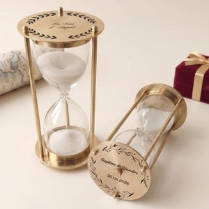May include: Two brass hourglasses with clear glass chambers filled with white sand. One hourglass is upright, the other is tilted. The top of the upright hourglass reads "La Tour d'Argent". The base of the tilted hourglass reads "Baptême de Léandre 30.05.2026".