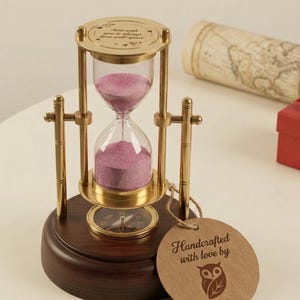May include: An ornate hourglass with a brass frame and a dark wooden base. The hourglass contains pink sand and has a compass design. A tag reads "Handcrafted with love by Owlauf."