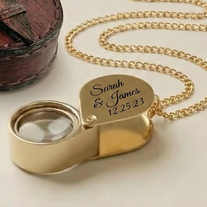 May include: A gold-colored magnifying glass pendant with a chain, engraved with "Sarah & James 12.25.23". A small, brown leather case and a red gift box are also in the image. A map is in the background.