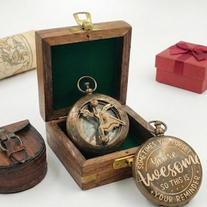 May include: A vintage brass compass with a sun and map design sits inside a wooden box with a green interior. A second compass with the text "Sometimes You Forget You're Awesome" is also present. A small red gift box is in the background.