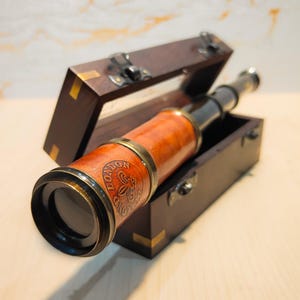 Puede incluir: Un telescopio de latón y cuero de estilo vintage, con la inscripción "LONDON". Se muestra dentro de una caja de madera oscura abierta, con detalles de latón, sobre una superficie de madera clara.