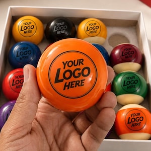 May include: A set of multi-colored billiard balls arranged in a white box, each featuring the text YOUR LOGO HERE. One bright orange ball with the same text is held in the foreground. The balls are glossy, round, and feature various solid colors.