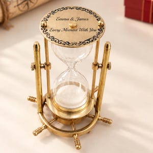 May include: A brass hourglass featuring a ship's wheel base and a top plate engraved with the text Emma & James and Every Moment With You. The hourglass contains white sand and is supported by two vertical brass pillars with side adjustment knobs.