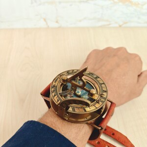 May include: A vintage brass sundial watch with a leather strap. The watch features Roman numerals and intricate detailing. The leather strap is a reddish-brown color. The watch is designed to be worn on the wrist.