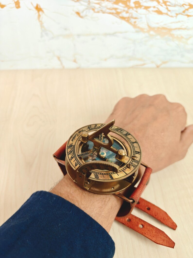 May include: A vintage brass sundial watch with a leather strap. The watch features Roman numerals and intricate detailing. The leather strap is a reddish-brown color. The watch is designed to be worn on the wrist.