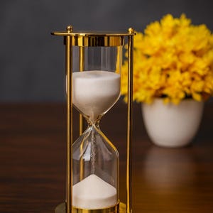 May include: A gold-colored hourglass with clear glass chambers filled with white sand. The hourglass has a decorative, ornate design. A bouquet of yellow flowers in a white vase is in the background, set on a dark wooden surface.