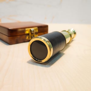 Peut inclure: Un télescope noir et doré avec un corps gainé de cuir et des accents en laiton. Le télescope est posé sur une surface en bois clair, avec une boîte en bois marron en arrière-plan.