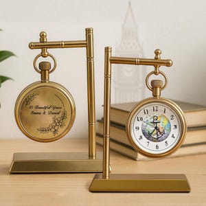 May include: Two gold-colored pocket watch displays. One watch has a white face with black numbers and an anchor design. The other watch has the text "10 Beautiful Years Emma & Daniel" with floral accents.