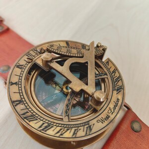 May include: A vintage brass compass with a leather strap. The compass features Roman numerals and the text "West London." The compass is a warm brass color, and the leather strap is a reddish-brown.