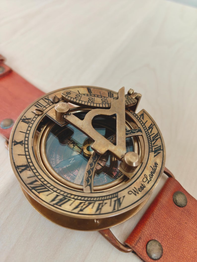 May include: A vintage brass compass with a leather strap. The compass features Roman numerals and the text "West London." The compass is a warm brass color, and the leather strap is a reddish-brown.