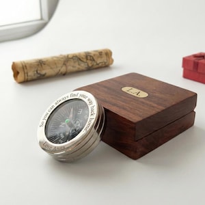 May include: A silver compass with the inscription "So you can always find your way back home" and "Love Anna." The compass sits next to a wooden box with the initials "L.A." engraved on a gold plate. A rolled-up map is in the background.