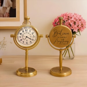 May include: Two gold-coloured, vintage-style desk clocks. One clock displays the time, while the other features the text "Believe in Yourself, Anything is Possible." Both clocks have a round face and stand on a decorative base.