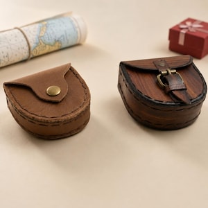 May include: Two small, semi-circular pouches made of leather. The left pouch is light brown with a simple brass snap closure. The right pouch is dark brown with a visible wood-grain texture, featuring a leather strap and a brass buckle closure.