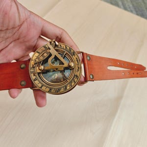 May include: A vintage-style brass sundial compass with a brown leather strap. The compass features Roman numerals and a detailed dial. The leather strap is secured with brass rivets. The compass is a navigational tool.