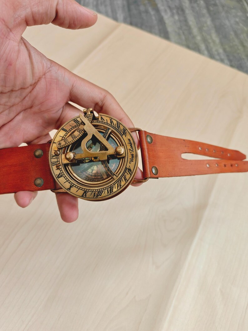 May include: A vintage-style brass sundial compass with a brown leather strap. The compass features Roman numerals and a detailed dial. The leather strap is secured with brass rivets. The compass is a navigational tool.