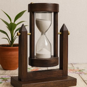 Puede incluir: Un reloj de arena de madera marrón oscuro con una cámara de vidrio transparente llena de arena blanca. El reloj de arena está sostenido por una base rectangular y postes verticales, con detalles en latón. Una planta en maceta está en el fondo.