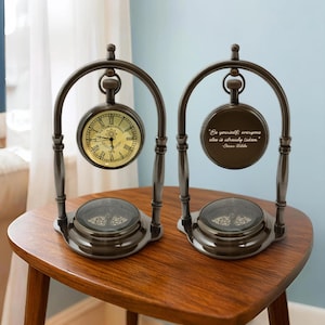 Puede incluir: Dos relojes de escritorio de estilo antiguo con acabado bronce. Uno muestra una esfera tradicional, el otro una cita de Oscar Wilde. Ambos tienen un diseño de brújula en la base y están sobre una mesa de madera.