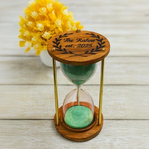 May include: Wooden hourglass with green sand, brass supports, and engraved text "The Rahns est. 2025". The hourglass has a clear glass centre and is set on a wooden base. A yellow flower arrangement is in the background.