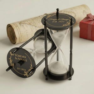 May include: Black hourglass with white sand, featuring the text "Forever & Always Est. 2024" in gold lettering. The hourglass is set against a backdrop of a rolled-up map and a red gift box, suggesting a theme of time and remembrance.