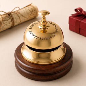 Puede incluir: Una campana de servicio de metal dorado pulido montada sobre una base circular de madera de color marrón oscuro. La campana presenta una inscripción grabada PERFECT ORDER AWARD 2026 SALES KICKOFF, con un pequeño pomo superior.