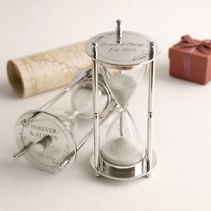 May include: Two silver hourglasses with clear glass chambers filled with white sand. The top and bottom of the hourglasses are engraved with "Forever & Always Est. 2024" and floral designs. A rolled-up map and a small gift box are in the background.