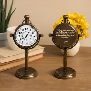 May include: Two antique-style brass clocks. One clock displays the time, while the other features the quote: "May your journey through time be filled with endless love and laughter." The clocks have a round face and stand on a decorative base.