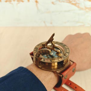 May include: A vintage brass sundial compass watch with a leather strap. The compass features a detailed dial and a gnomon. The watch is secured with a reddish-brown leather strap. The watch is approximately 2 inches in diameter.