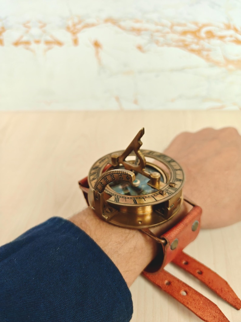 May include: A vintage brass sundial compass watch with a leather strap. The compass features a detailed dial and a gnomon. The watch is secured with a reddish-brown leather strap. The watch is approximately 2 inches in diameter.