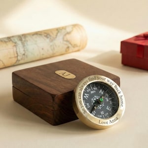 May include: A dark wooden box with a gold plate marked "AJ" next to a gold compass. The compass has a black face with white text that reads "So you can always find your way back home. Love Anna."