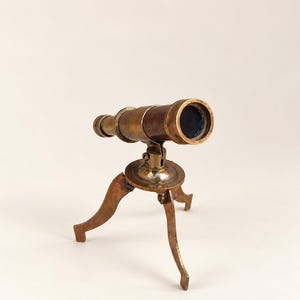 Peut inclure: Un télescope en laiton de style ancien monté sur un trépied à trois pieds. Le télescope présente un corps cylindrique avec une finition sombre et patinée ainsi qu'une ouverture de lentille circulaire, soutenu par un mécanisme pivotant central.